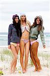 Three attractive young women in bikinis are standing on the beach.  They are also wearing sweaters, a coat and knit caps. Vertical shot.