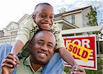 Happy African American Father and Son in Front of New Home and Real Estate Sign.