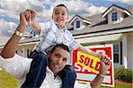 Excited Hispanic Father and Son with Sold For Sale Real Estate Sign in Front of House.