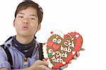 Chinese man, dressed with Bavarian Lederhose holds an Oktoberfest gingerbread heart in camera and gives a kiss. Isolated on white.