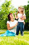 happy young mother sitting in park with her beautiful daughter blowing soap bubbles