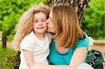happy young mother sitting in park under tree and kissing her smiling small daughter