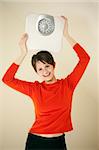 Attractive young woman holds a bathroom scale above her head while smiling at the camera. Vertical shot.