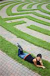 Beautiful young Asian woman lies down on the grass of a park labyrinth while enjoying a conversation on her cellphone. Vertical shot.