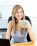 Glowing businesswoman holding a cup using her laptop at her desk