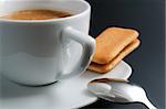 White porcelain cup of freshly brewed coffee close-up arranged with sandwich-biscuit spoon and plate on dark background