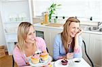Two cheerful female friends eating pastries and drinking coffee in the kitchen at home