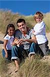 A man mixed race girl and young boy, father, son and daughter, sitting down and having fun in the sand dunes of a sunny beach