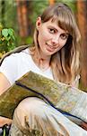 beautiful young woman sitting on  forest floor with backpack and map, smiling and looking in camera