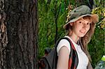 beautiful young woman standing near the tree in forest with backpack and bottle of water, smiling and looking in camera