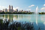 New York City Central Park fountain and urban Manhattan skyline with skyscrapers and trees lake reflection with blue sky and white cloud.