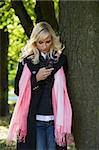 young blond woman with hairstyle in winter dress using her telephone near a tree in a forest