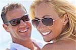A happy and attractive man and woman couple wearing sunglasses and smiling in sunshine at the beach