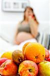 Fruit platter on table and pregnant woman sitting on sofa at home in background
