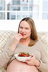 Cute pregnant woman eating a strawberry sitting at home