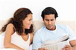 Happy woman looking at her serious boyfriend reading the newspaper on the bed