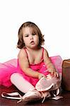 Little girl wearing a pink ballet outfit sitting next to an antique suitcase trying on oversized ballet shoes