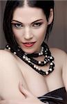 Sexy naked young caucasian adult woman with red lips, short black hair and a pierced eyebrow, covered in a dark satin sheet and wearing a black and white pearl string necklace