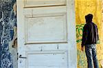 Hooded figure in abandoned house. Empty room weathered walls torn wallpaper.