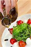 Pouring olive oil salad dressing onto tomato, mozarella and rocket salad with basil garnish and balsamic vinegar