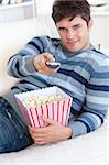 Relaxed young man eating popcorn and holding a remote lying on the sofa at home