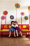 Caucasian and hispanic female preschoolers sitting on bench in kindergarten, playing and smiling. Vertical shape, full length, front view