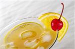 Maraschino cherry and orange slice on the rim of a cocktail glass with ice cubes, orange juice and orange slice inside photographed on white sand (Selective Focus, Focus on the cherry and the ice cubes in the front on the right of the glass)