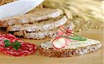 Rolled salami slice and cream cheese with chives on half a slice of brown bread on wooden board and ingredients (Selective Focus, Focus on the roll and bread slice in the front and the front of salami slices on the board)