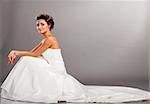 happy beautiful bride is sitting in wedding dress and looking at camera on grey background