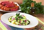 Fresh potato salad with cucumber, red and green onion made with an oil dressing with barbecued sausages, oil and parsley in the back (Selective Focus, Focus on the parsley leaf on the salad and its surroundings)