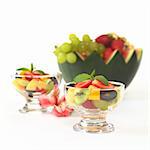Fresh fruit salad with strawberry, kiwi, mango and grapes in glass bowl garnished with mint leaf with Inca Lily and a watermelon bowl full of fruits in the back (Selective Focus, Focus on the mint leaf in the first bowl)