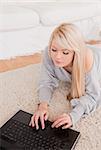 Pretty young blond woman relaxing on laptop lying on a carpet in the living room