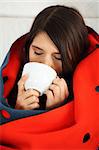 Young woman caught cold, wrapped up in blanket, drinking something hot from cup.