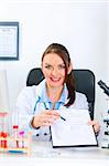 Smiling medical doctor woman sitting at office table with document and pen for signing