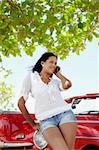 young adult brunette woman leaning on convertible red car and talking on mobile phone. Vertical shape, front view, three quarter length