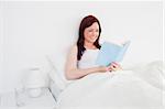 Beautiful red-haired woman reading a book while sitting on her bed