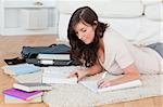 Young brunette female writing on a notebook while lying on a carpet in the living room