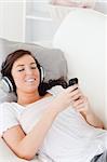 Charming brunette woman writing a text on her mobile while lying on a sofa in the living room