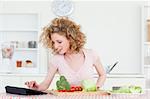Beautiful blonde woman relaxing with her tablet while cooking some vegetables in the kitchen in her appartment