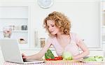 Pretty blonde woman relaxing with her laptop while cooking some vegetables in the kitchen in her appartment