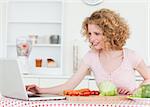 Attractive blonde woman relaxing with her laptop while cooking some vegetables in the kitchen in her appartment