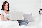 Close up of a charming brunette sitting on a sofa using a laptop looking at the camera