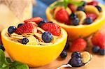 Wholewheat cereal with fresh strawberries and blueberries served in a half orange bowl with yogurt and fruit salad in the back (Selective Focus, Focus on the blueberry on the right front of the cereal)