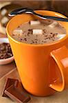 Hot chocolate with marshmallows in orange cup with a teaspoon on the rim and chocolate pieces on the side (Selective Focus, Focus on the marshmallows in the middle of the hot chocolate)