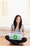 A sitting woman is holding a recycling bin in her living room