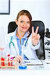 Smiling medical doctor woman sitting at office table and showing victory gesture