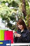 Junge Frau mit Einkaufstüten draußen sitzen und betrachten Telefon