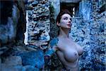 Blue-toned portrait of tattooed sexy nude Caucasian woman next to concrete wall.