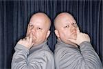 Caucasian bald identical twin men standing back to back and looking at viewer with hands to mouth.