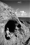 Young nude Asian woman mediatating in cave with coastline of Maui, Hawaii.